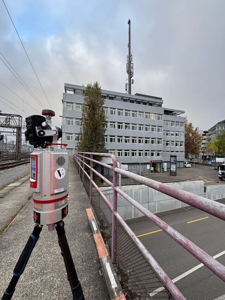 Vermessungsausrüstung auf einem Stativ neben Eisenbahnschienen, mit moderner Bürogebäude im Hintergrund.