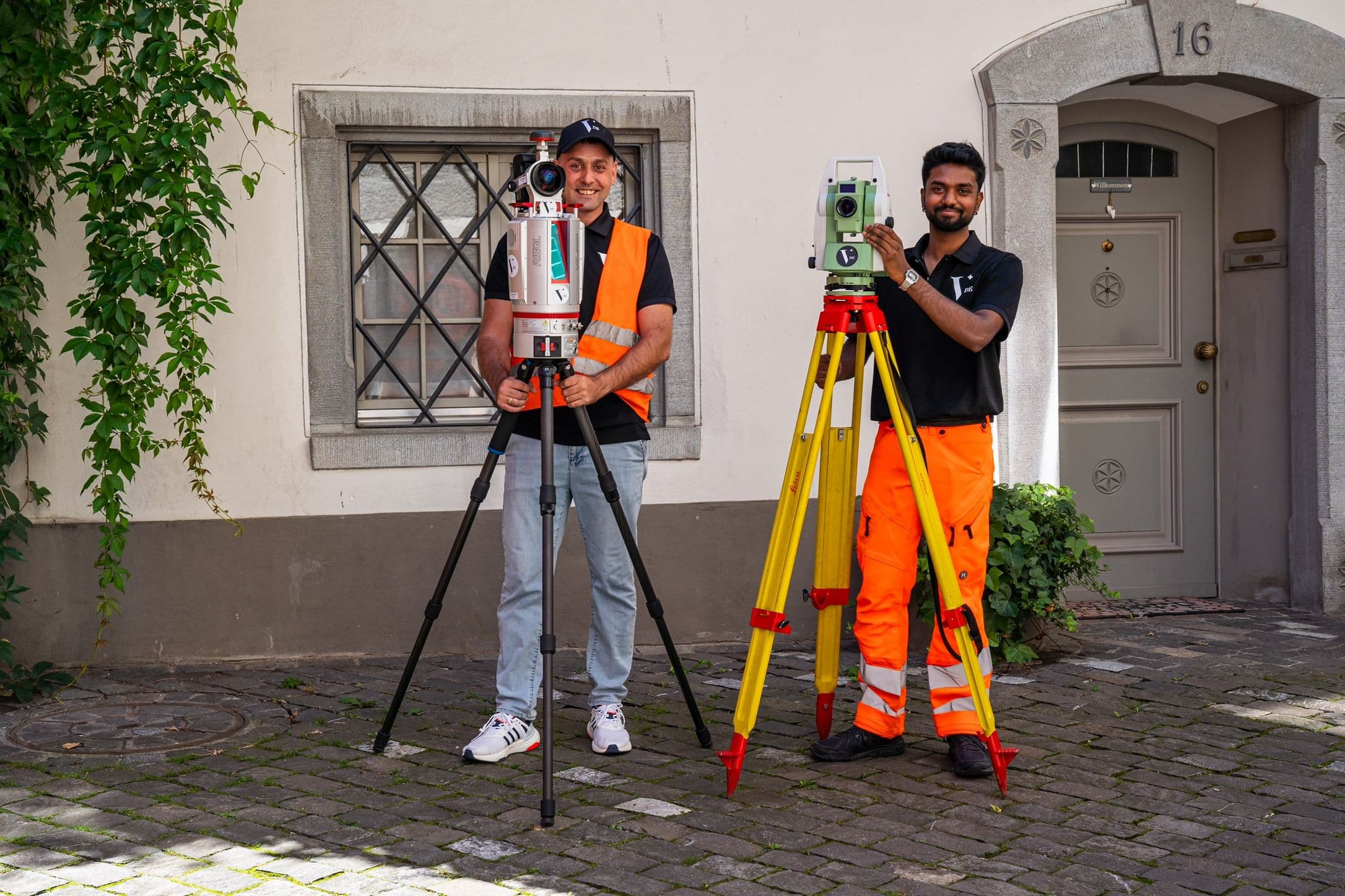 Zwei Vermessungstechniker mit Theodoliten vor einem Haus.